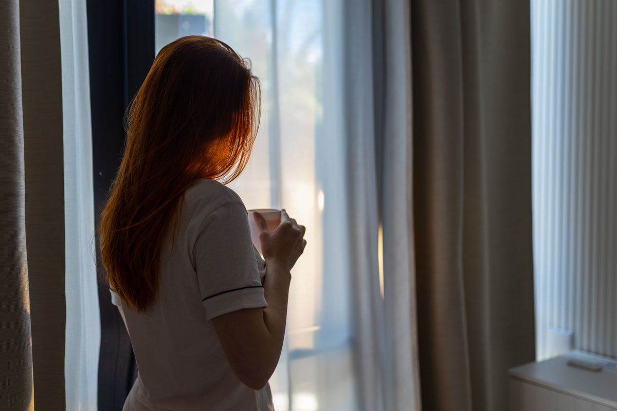 A young woman holding a coffee cup, looking through a window in warm morning light